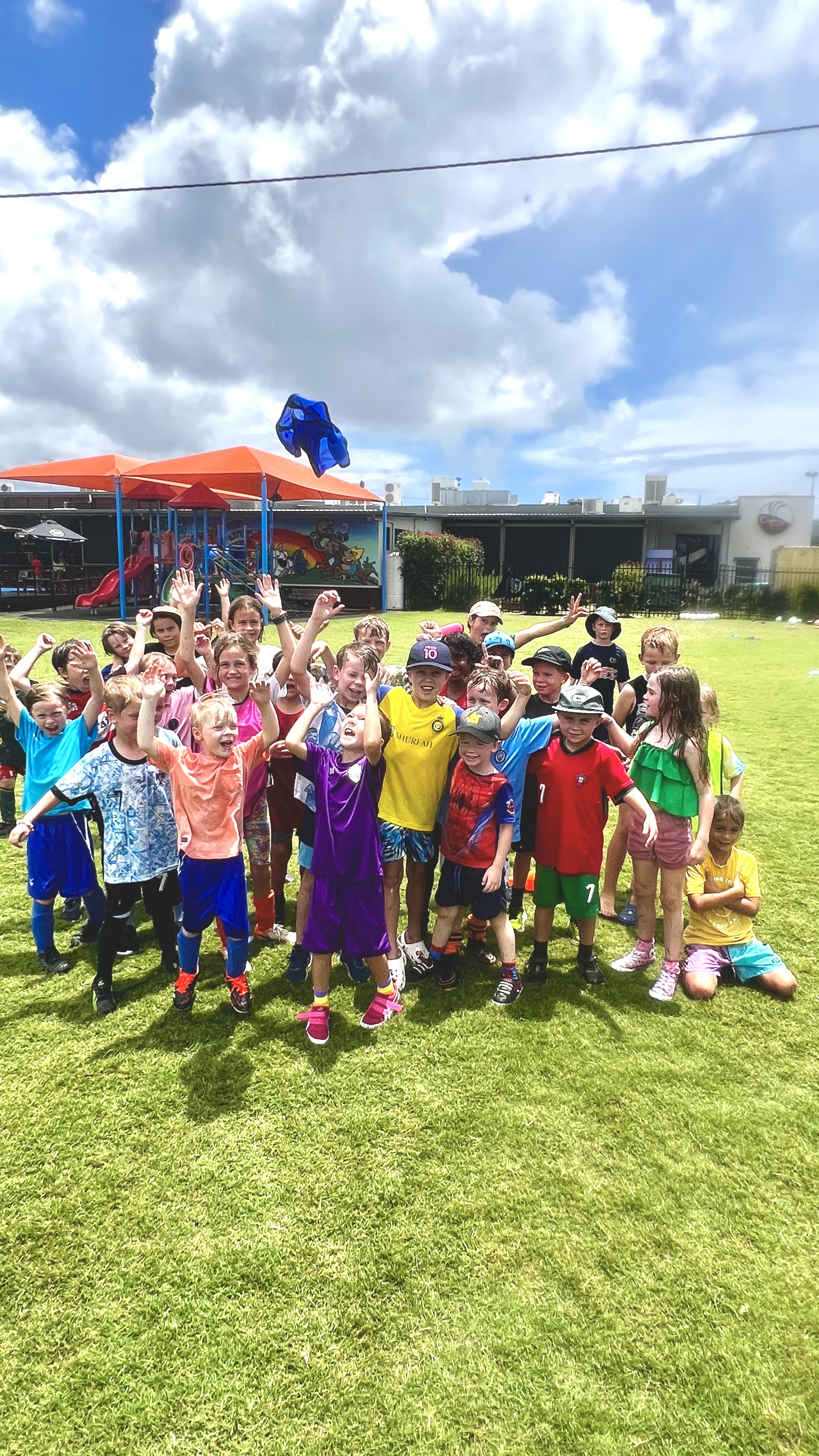 A large group of excited children in colourful sports clothes cheer together on a sunny field.